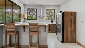A modern kitchen with a central island, wooden chairs, a stainless steel refrigerator, and large windows. - Stylish 3-Bedroom Tropical Escape with Pool & Outdoor Living, 1 Block from Playa Potrero (Playa Potrero)