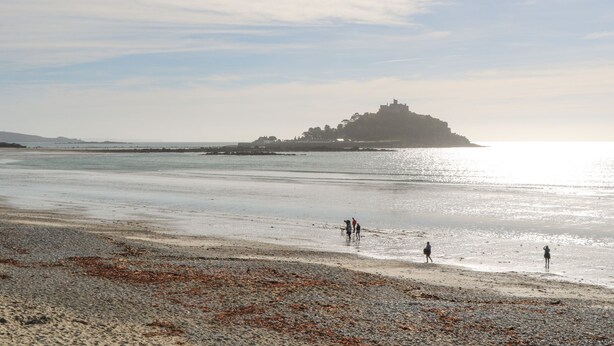 A beach with a distant castle-like structure, calm water, and a few people walking along the shore. - Scillonia (Penzance)
