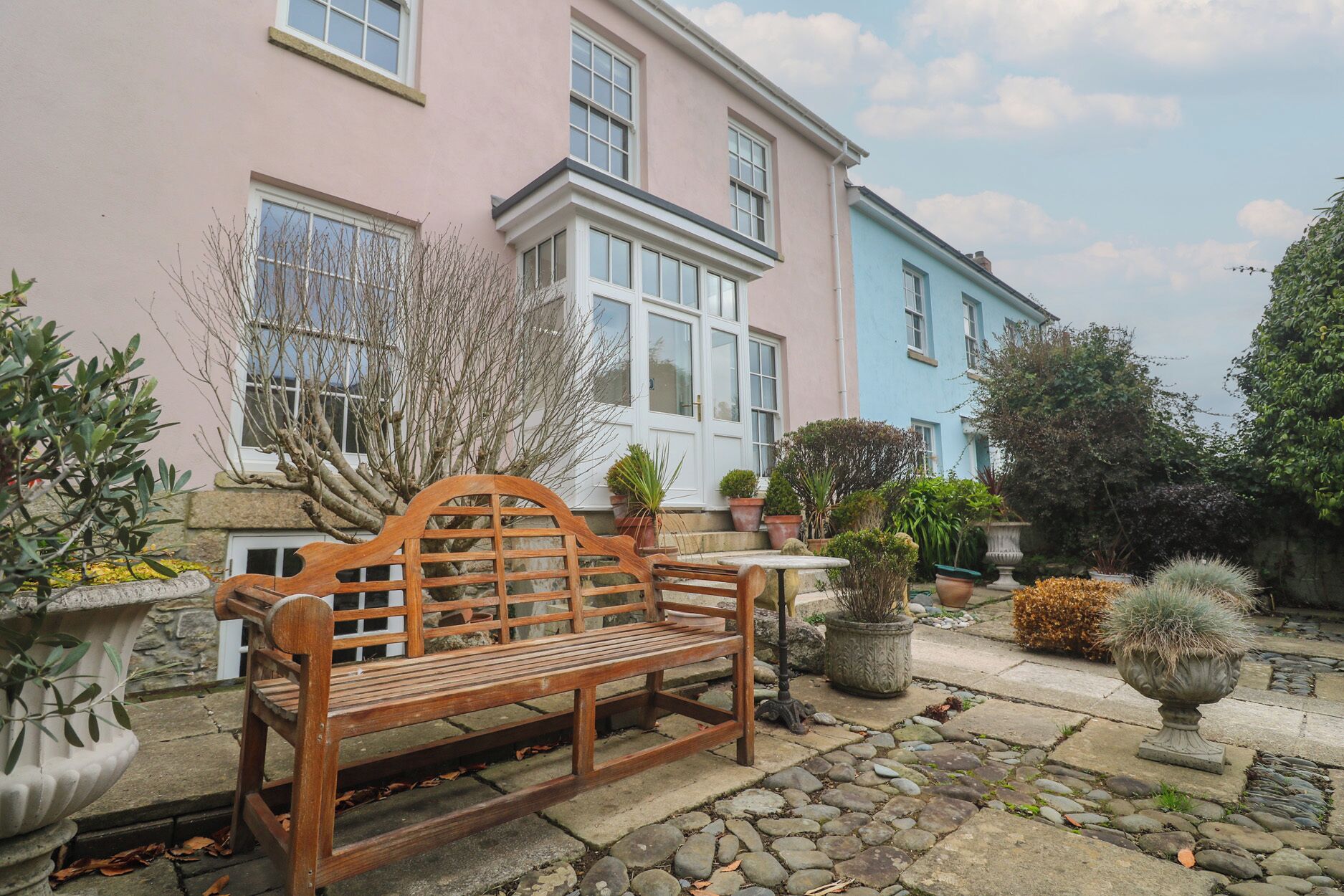 A wooden bench in a garden with a stone pathway, potted plants, and a pink and blue house in the background.