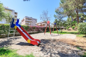 A playground with a slide and swings, surrounded by greenery and buildings. - Ground floor duplex large terrace UHC GOLDEN PINEDA APARTMENTS 206 (La Pineda)