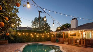 Una piscina con muelle de madera, rodeada por un seto y luces de cuerda.
