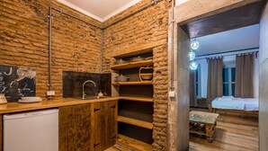 Eine kleine Küche mit Holzausbau, Spüle und kleinem Kühlschrank. Ein Holzregal beherbergt Geschirr und einen Korb. Eine Holzkonstruktion ist in der Ecke sichtbar. Der Raum verfügt über eine rustikale Backsteinwand und Holzboden.