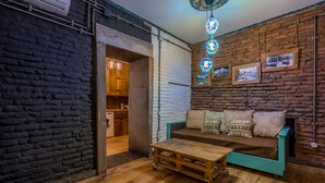 Ein gemütliches Zimmer mit Holzboden, einem türkisfarbenen Sofa mit gemusterten Kissen, einem Holztisch und einer Ziegelwand mit gerahmten Bildern.