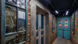 Ein schmaler Korridor mit einer rustikalen Ziegelwand, einer Holztür mit blauem Griff und einem farbenfrohen Buntglasfenster.