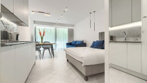 A modern hotel room with a bed, a sofa, a dining area with a table and chairs, and a kitchen area.