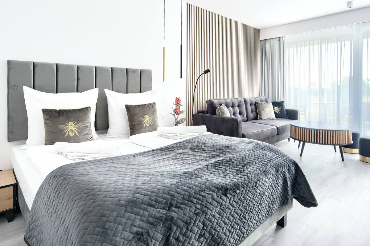 Een moderne hotelkamer met een groot bed, een grijze gestoffeerde bank, een ronde salontafel en uitzicht op het groen door grote ramen.