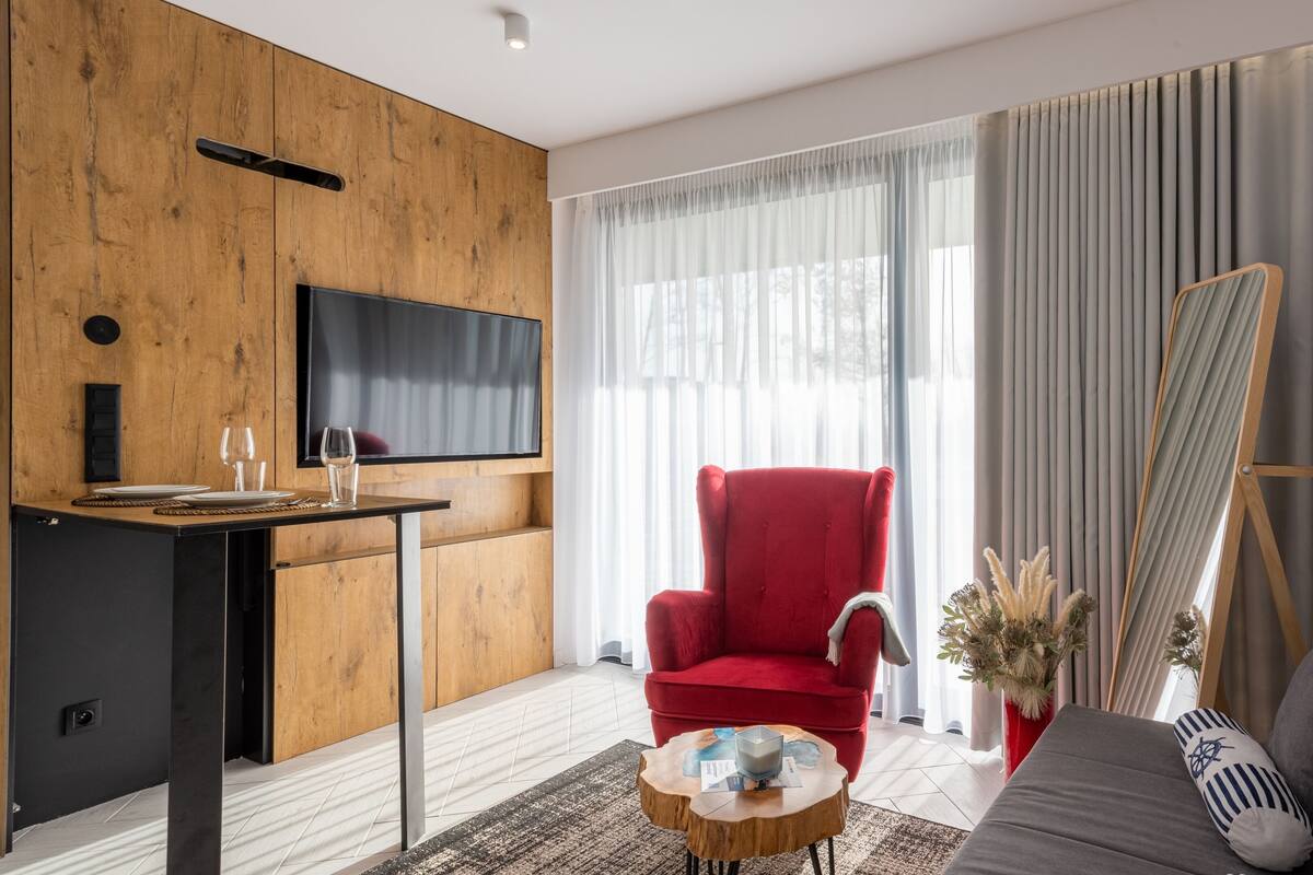 Een moderne woonkamer met een houten accentmuur, een rode fauteuil, een grijze bank, een flatscreen-tv aan de muur en een groot raam met transparante gordijnen.