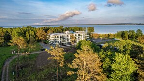A modern multi-story building with a flat roof, surrounded by dense greenery and a body of water in the background.