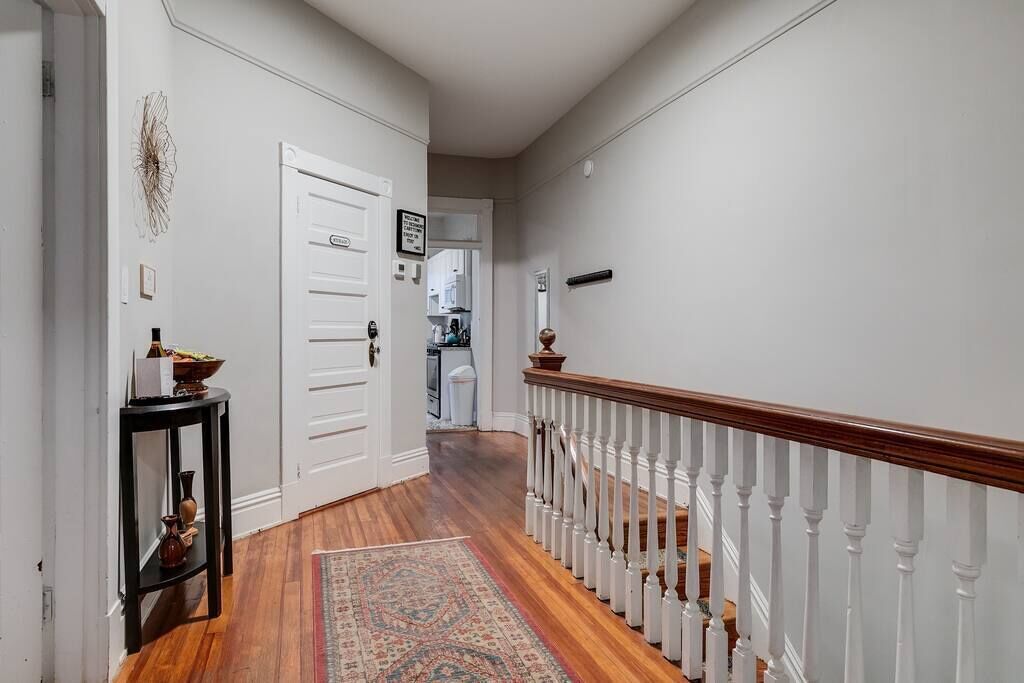 Un couloir doté d’un plancher en bois, d’une porte blanche, d’une petite table ornée d’objets décoratifs et d’un escalier avec une rampe blanche.