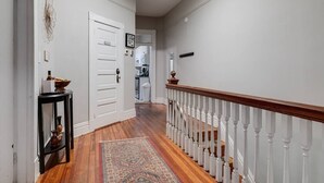 Un pasillo con suelo de madera, una puerta blanca, una mesita con adornos y una escalera con barandilla blanca.