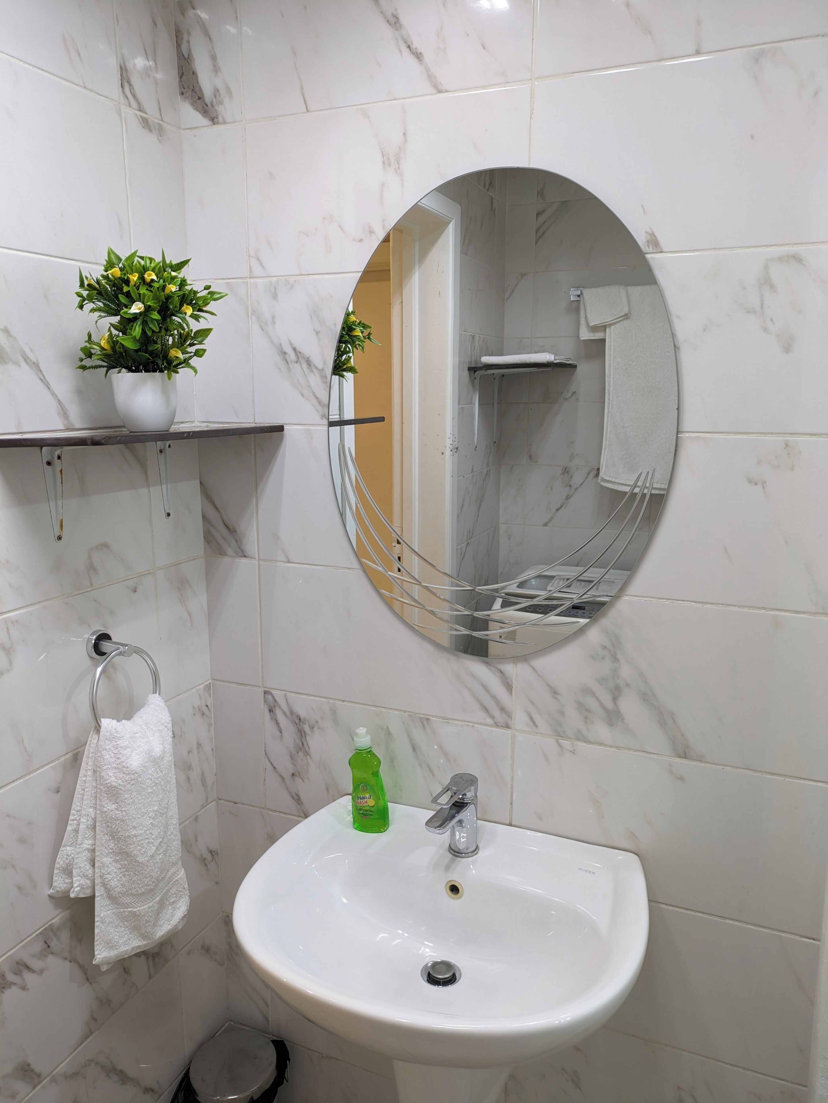 A bathroom with a white marble sink, a round mirror, a towel rack, and a potted plant.