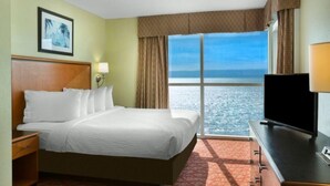 A hotel room with a large bed, a television, a desk, and a window with a view of the sea. - Ocean's Edge Bliss with Dual Balconies (Myrtle Beach)