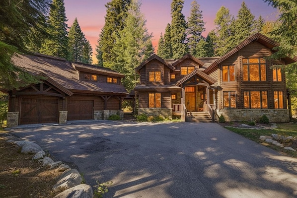 A large, multi-story log cabin with a stone foundation, a driveway, and a forested backdrop.