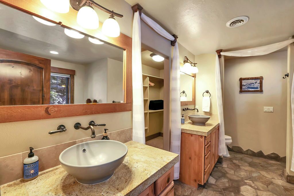 A bathroom with a large vessel sink, wooden cabinets, a mirror with lights, and a stone-patterned floor.