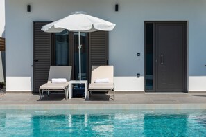 Un área junto a la piscina con dos sillones de descanso y una sombrilla blanca, una puerta y una ventana con persianas.
