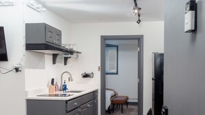 Une cuisine compacte avec des meubles gris, un évier et un coin repas comprenant une table et un banc. Une porte donne accès à une chambre à coucher avec un lit et une petite table.