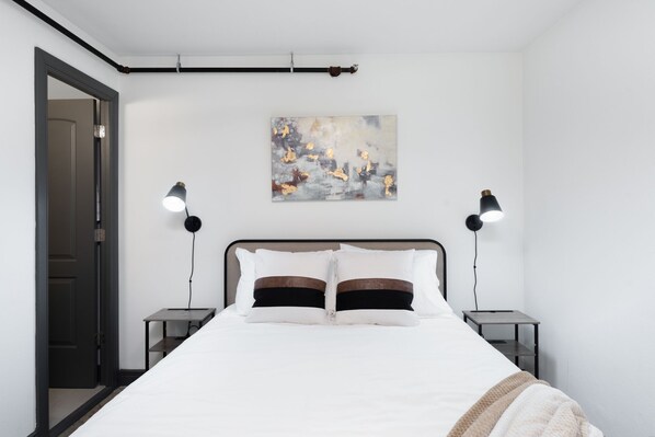 A bedroom with a bed, two bedside tables, a nightstand, a painting on the wall, and a door.