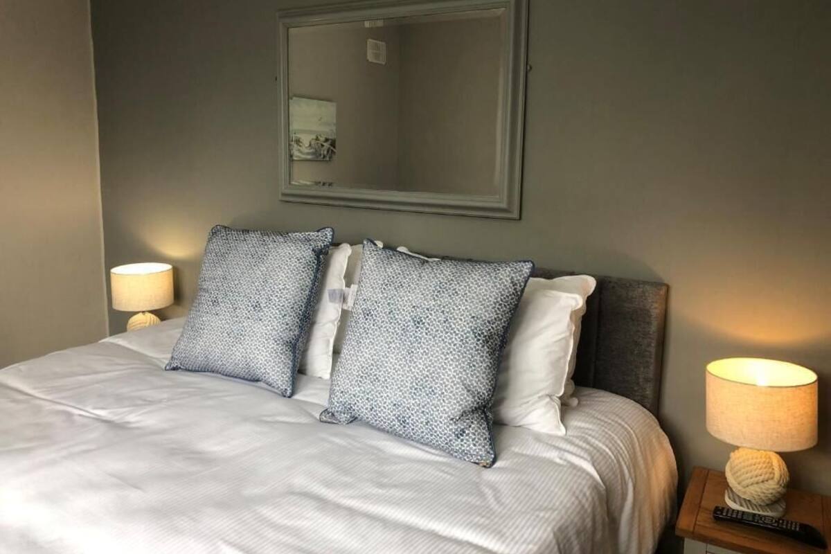 Una cama bien hecha con almohadas estampadas, una lámpara de noche y un espejo enmarcado en la pared.