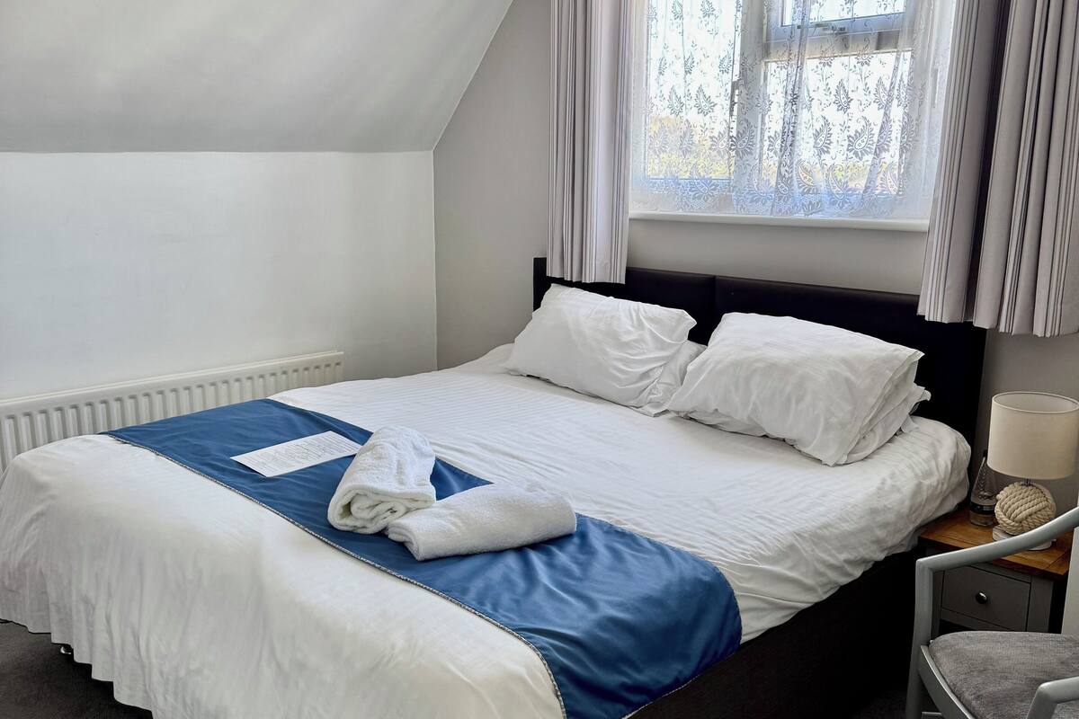 Una cama bien hecha con ropa de cama blanca y azul, una lámpara de noche y una silla.