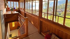 A wooden interior with a staircase leading to a loft area, large windows offering a view of a valley, and a cozy seating area with a red patterned cushion. - Wohnung Lake View (Schwanden bei Brienz)
