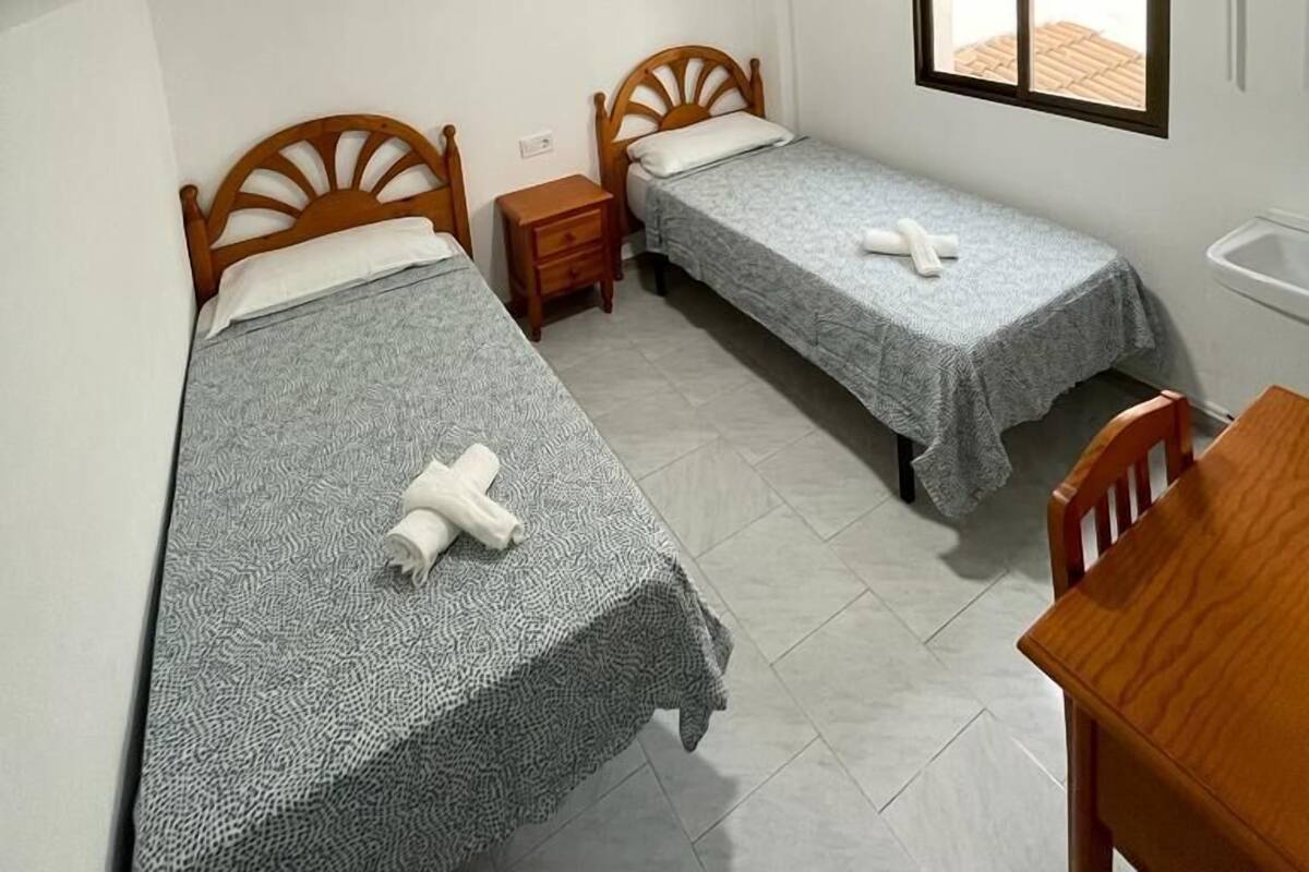 A small, clean room with two single beds, a wooden nightstand, a desk, and a window with a blind.