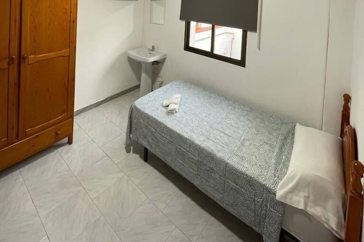 A small, clean room with a single bed, a wooden wardrobe, a sink, and a window with a blind.