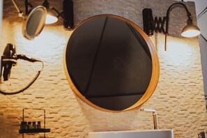 Un miroir rond, encadré de bois, est fixé sur un mur en briques, et deux lampes murales se trouvent de part et d’autre.