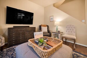 A living room with a flat-screen TV, a dark wooden dresser, a wicker ottoman, a cushioned chair, a small side table with a lamp, and a 'WELCOME' sign. - Mesquite 2BR Townhome with Pool & Gym Access (Mesquite)