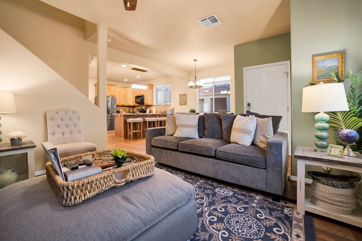 A modern living room with a grey sofa, a coffee table with books and a laptop, a patterned rug, and a kitchen in the background.