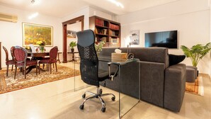 A modern living room with a glass coffee table, a dark grey sofa, a black office chair, a dining area with a wooden table and red upholstered chairs, a bookshelf, and a flat-screen TV mounted on the wall.
