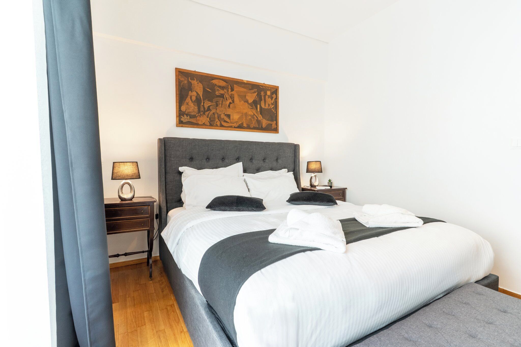A neatly made bed with white and grey bedding, two bedside lamps, and a large artwork above the bed.