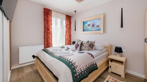 Quarto com cama de madeira, mesa de cabeceira com abajur, radiador e janela com cortinas.