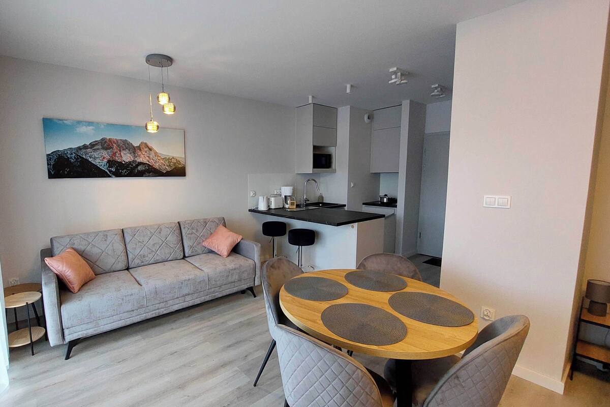 Una sala moderna con un sofá gris, una mesa de comedor redonda de madera y sillas grises, una cocina con encimera negra, y un cuadro de montañas en la pared.