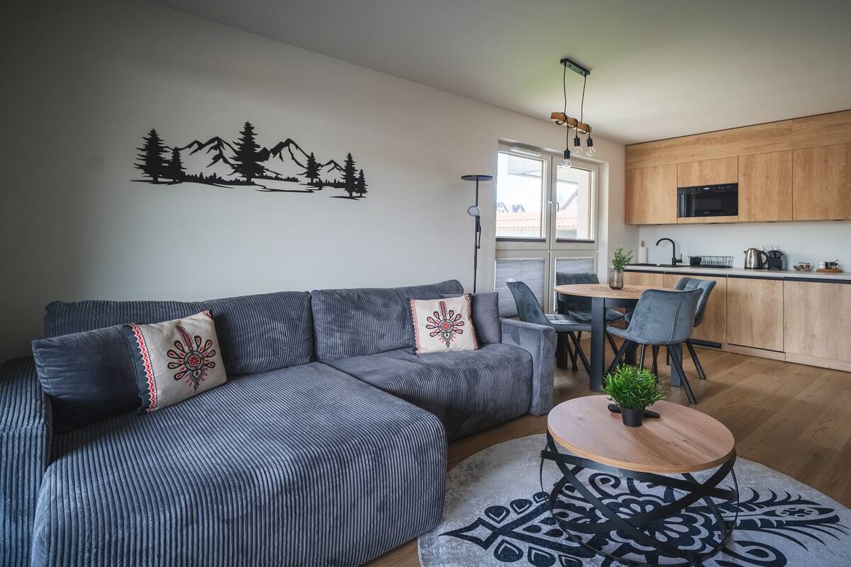 Una sala de estar moderna con un sofá gris, una mesa redonda y un comedor con mesa y sillas de madera.