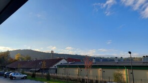 Vista de um edifício com telhado verde, um estacionamento com carros e uma colina arborizada ao fundo.