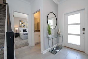 A modern interior with a staircase, a hallway leading to a bedroom, a glass console table with a mirror, and a door with a window.