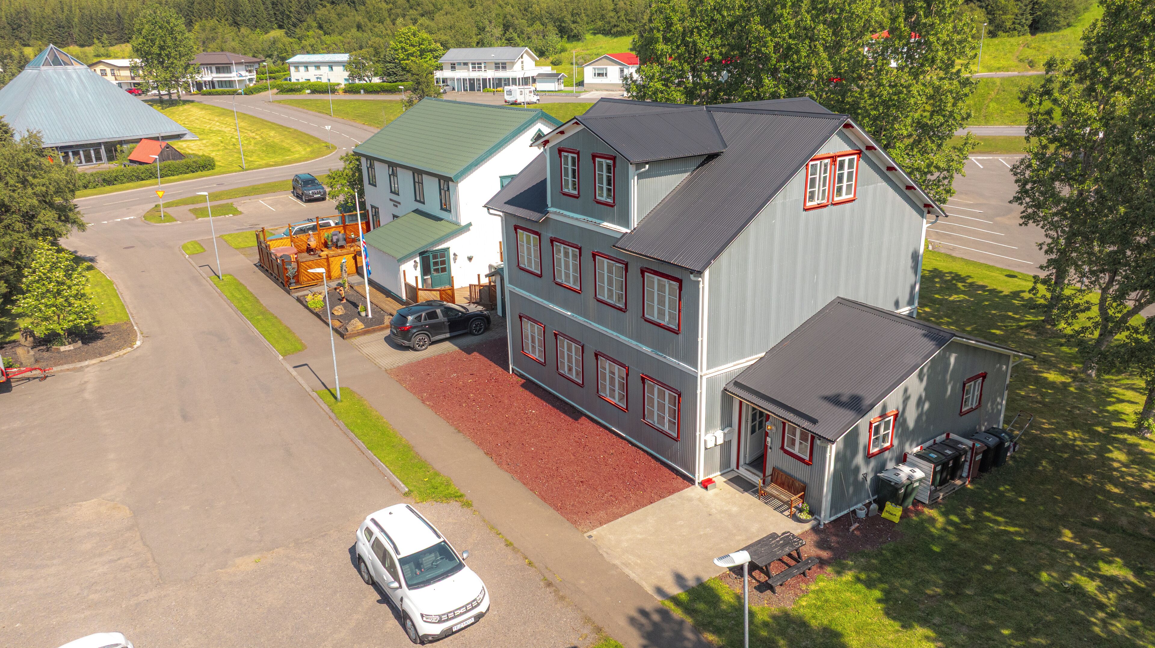A large two-story building with a grey exterior and red trim, a parking lot, and a small adjacent structure with a red roof.