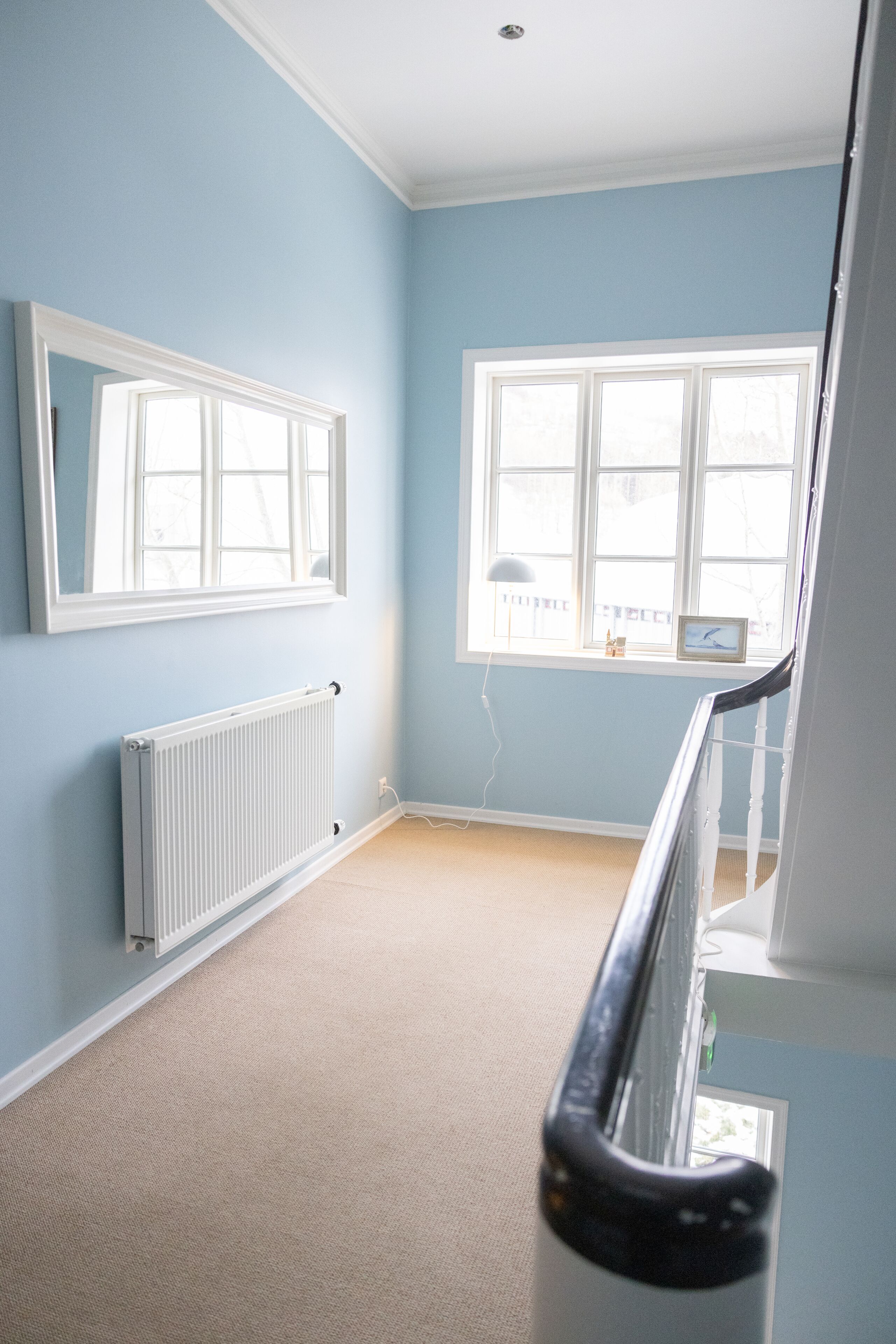 Une pièce aux murs bleu clair, avec un radiateur blanc, une grande fenêtre à cadre blanc et un plafond blanc.