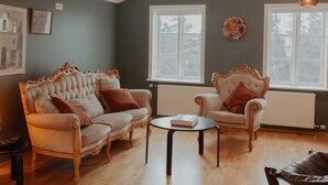 A living room with a high ceiling, two ornate armchairs, a small table, and a radiator.