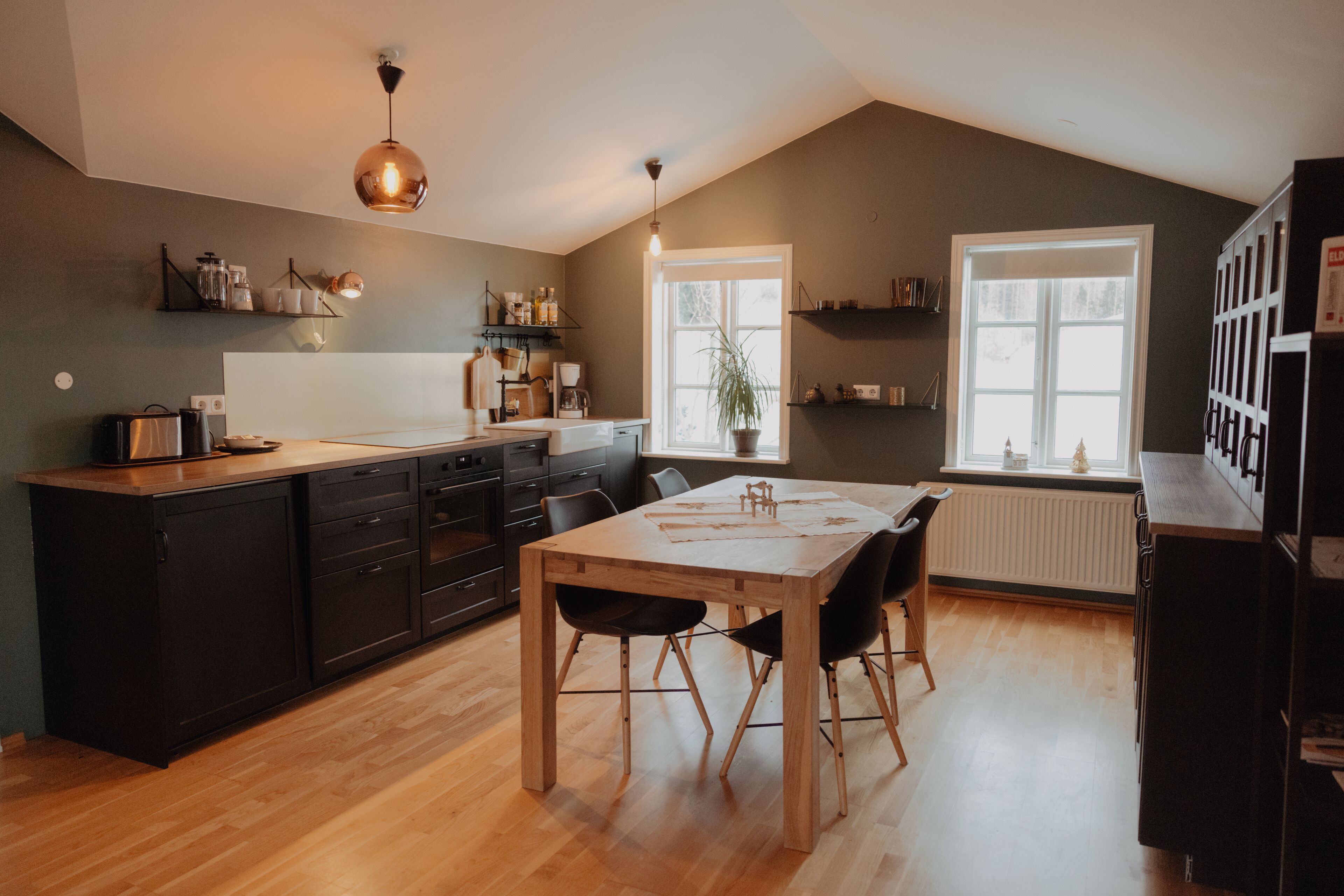 A modern kitchen with wooden cabinets, a dining table, and black chairs. There is a window providing natural light and a potted plant on the windowsill.