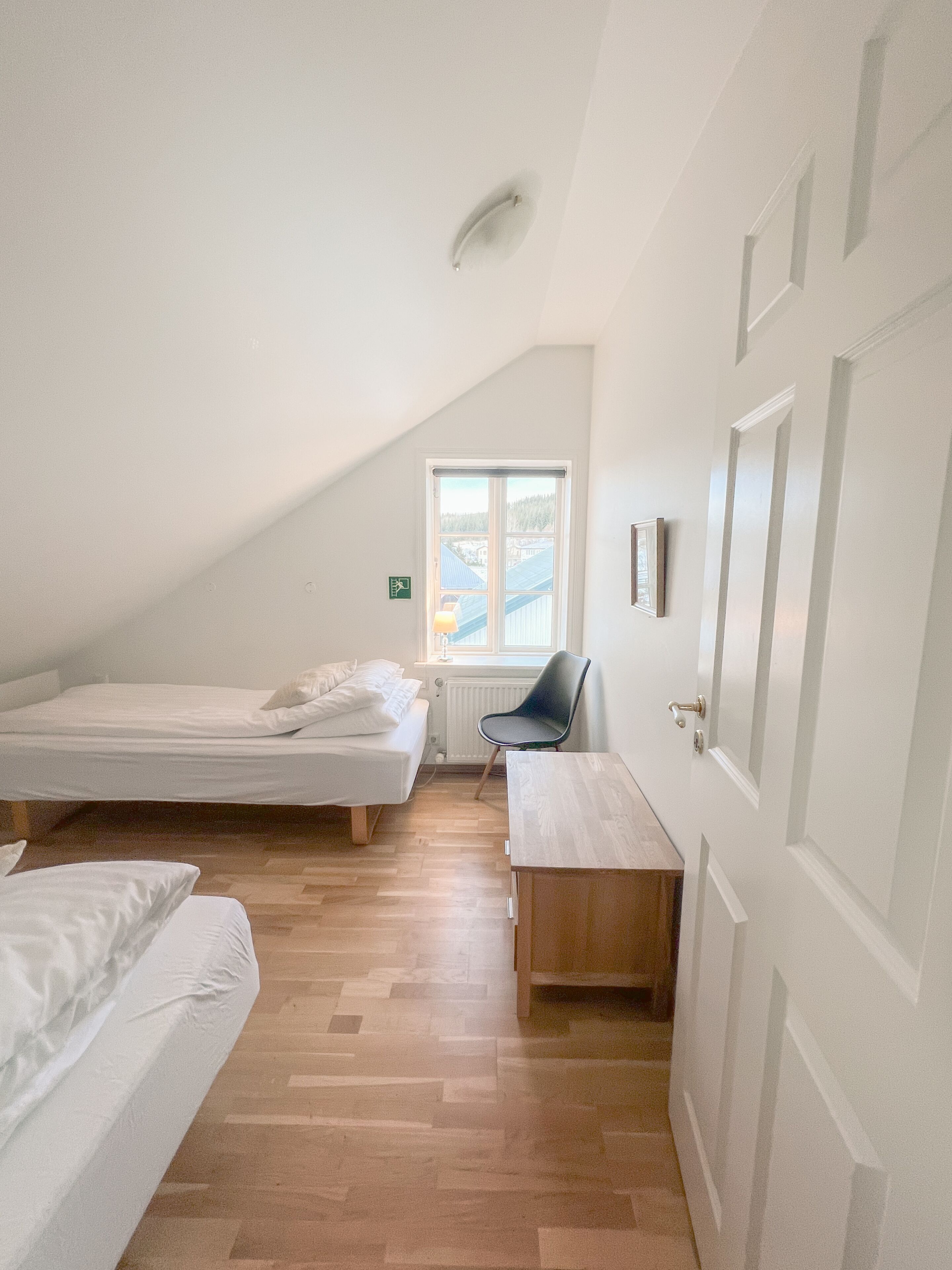 Ein Zimmer mit zwei Betten, einem Holztisch, einem Stuhl, einem Heizkörper und einem Fenster mit Blick auf Gebäude und Bäume.
