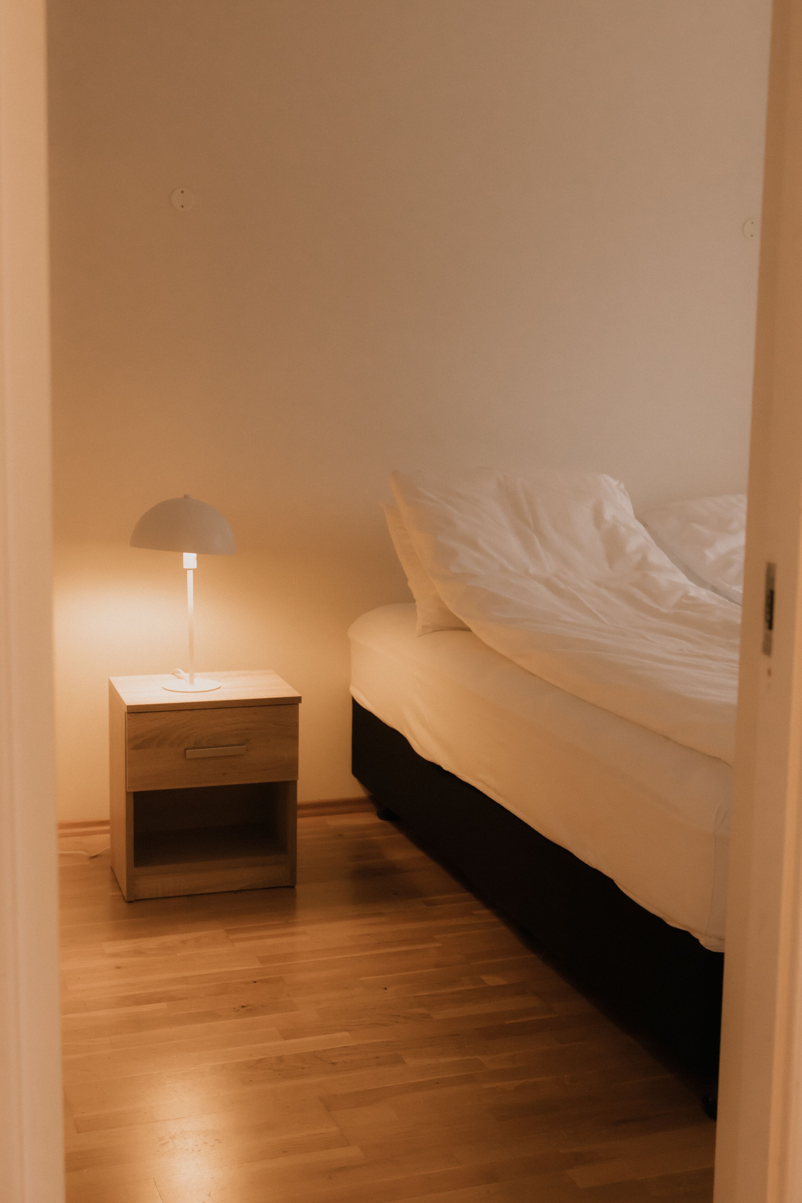 Un dormitorio con mesita de noche de madera, lámpara y cama con ropa blanca.