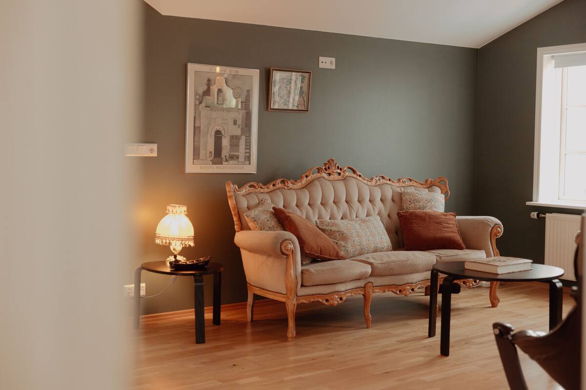 A living room with a tufted sofa, a small table, and a lamp. There are two framed pictures on the wall.