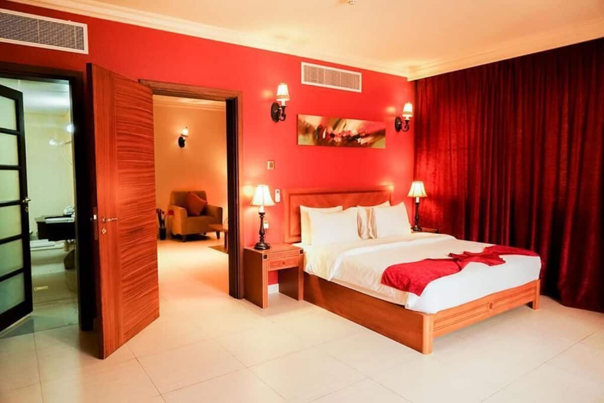 Une chambre d’hôtel avec un mur rouge, un lit en bois, deux lampes de chevet et une porte donnant sur une autre pièce.