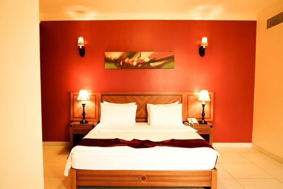 Une chambre d’hôtel avec un lit en bois, deux lampes de chevet, un tableau au mur et un mur d’accent de couleur rouge.