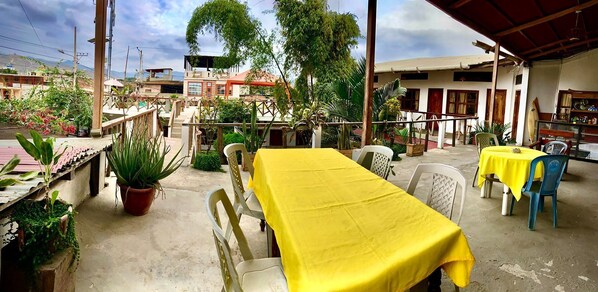 An outdoor patio with a yellow table and chairs, surrounded by potted plants and a view of buildings in the background.