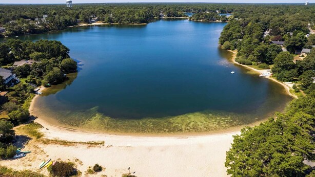 Vista aérea de un lago rodeado de bosque frondoso, con una playa de arena y botes.