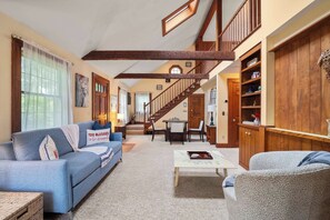 Ein geräumiges Wohnzimmer mit hoher Decke, freiliegenden Holzbalken, einer Treppe und einer gemütlichen Sitzecke mit Sofa und Sesseln.