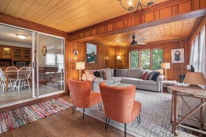 A cozy cabin interior with a dining area, a living room with a sofa and two chairs, and a large window providing natural light.