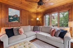 A cozy living room with a sectional sofa, a ceiling fan, a painting of a coastal scene, and large windows offering a view of greenery. - Walk to Private Beach on Buck's Pond! (Harwich)